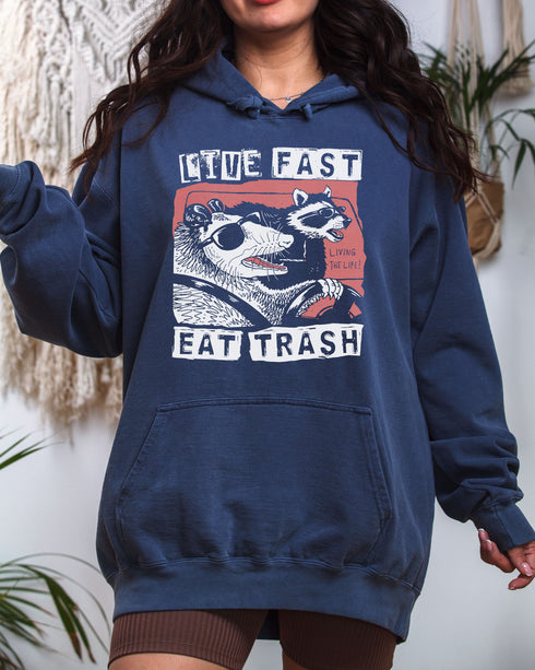 Woman wearing a denim-colored "Live Fast Eat Trash" hoodie with a retro graphic of a raccoon and opossum driving a car.